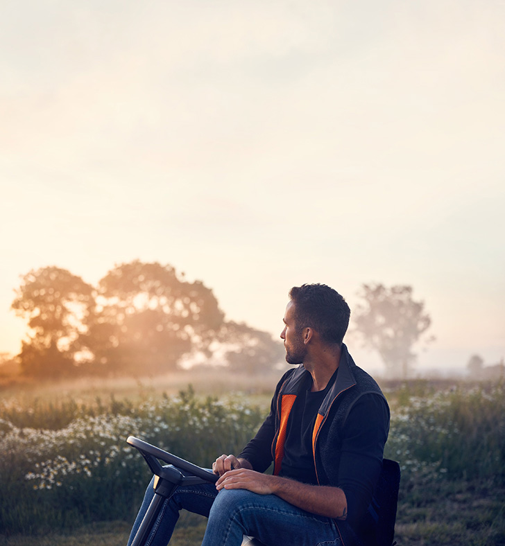 Un utilisateur profite d’un moment calme au lever du soleil avec un rider HUSQVARNA, conçu pour une tonte précise et confortable des grandes pelouses.