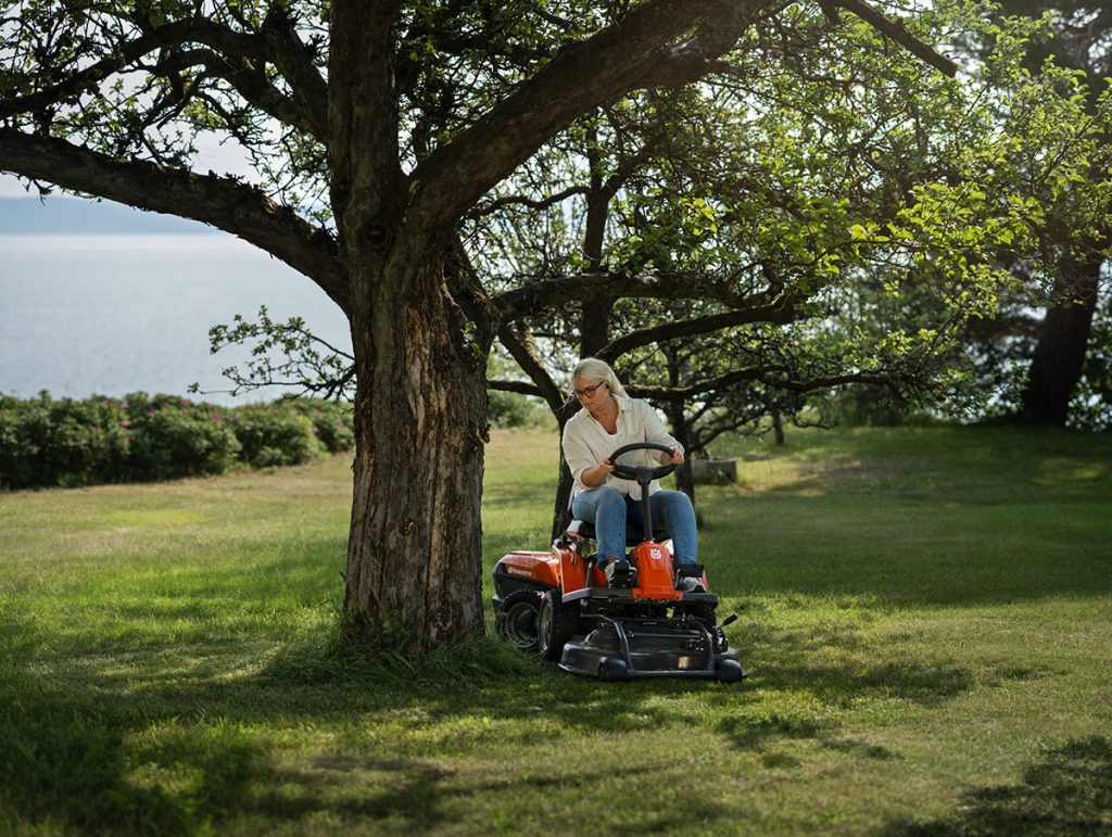 Utilisateur conduisant un rider HUSQVARNA sous un arbre dans un grand jardin pour tondre la pelouse avec une tondeuse autoportée.
