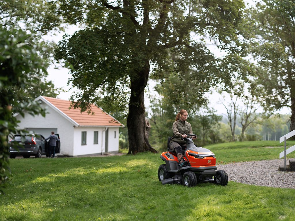 Utilisatrice conduisant un rider HUSQVARNA pour tondre la pelouse d’un grand jardin près d’une maison.