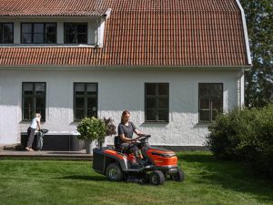 Utilisatrice tondant la pelouse avec un rider HUSQVARNA dans le jardin d’une maison.